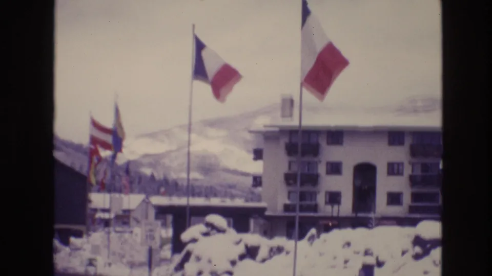Vail, Colorado 1974 frame showing several national flags, including a French tricolor, in front of a snow-surrounded hotel building and pale mountain backdrop.