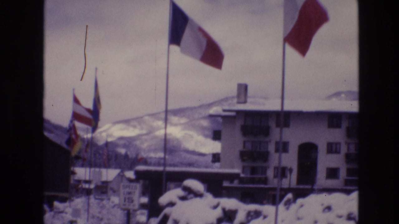 In Vail, Colorado, a 1974 8mm home-movie frame shows international flags, including a French flag, waving above snowbanks near a multi-story lodge and snowy hills.