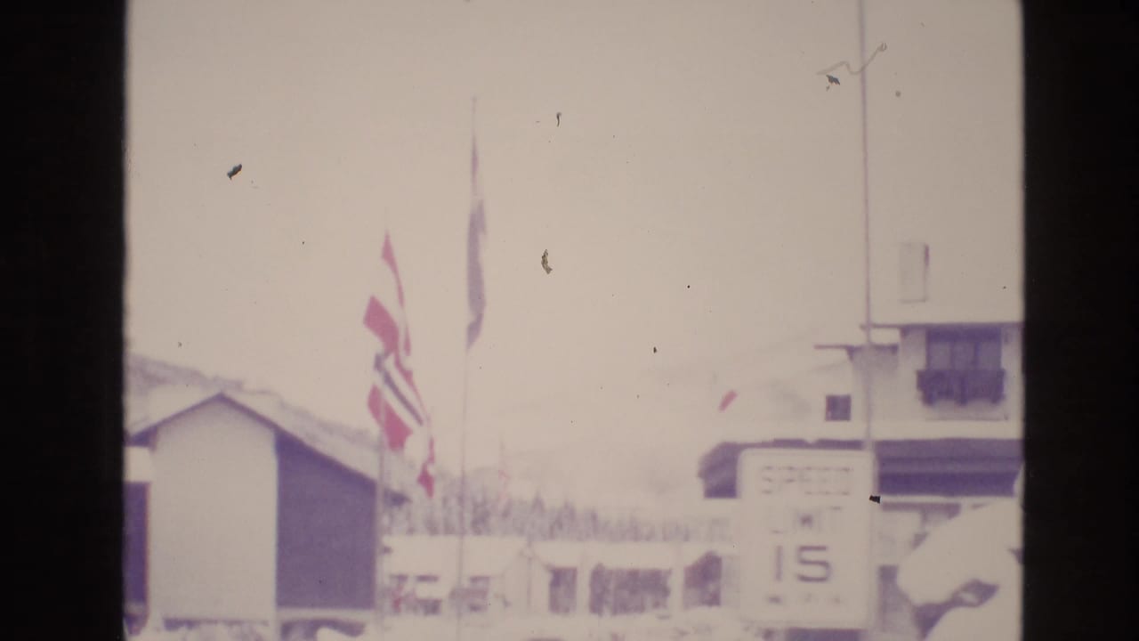 Overexposed snowy resort view with flags, a small building, and a speed limit sign.