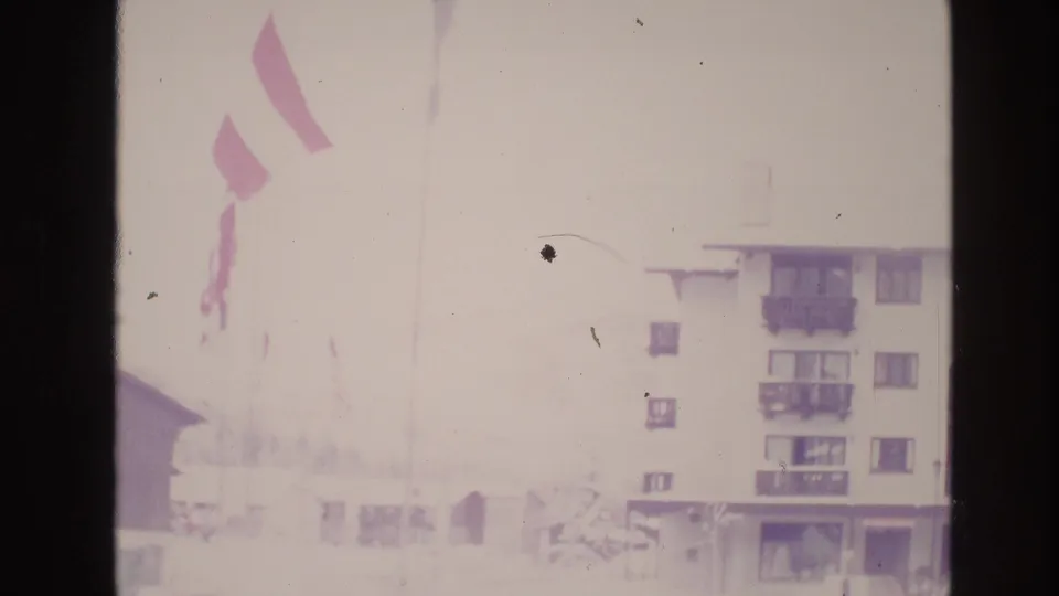 The 1974 Vail home-movie frame shows a high-exposure snowy lodge area with several flags and part of a multi-story resort building visible through faded color.