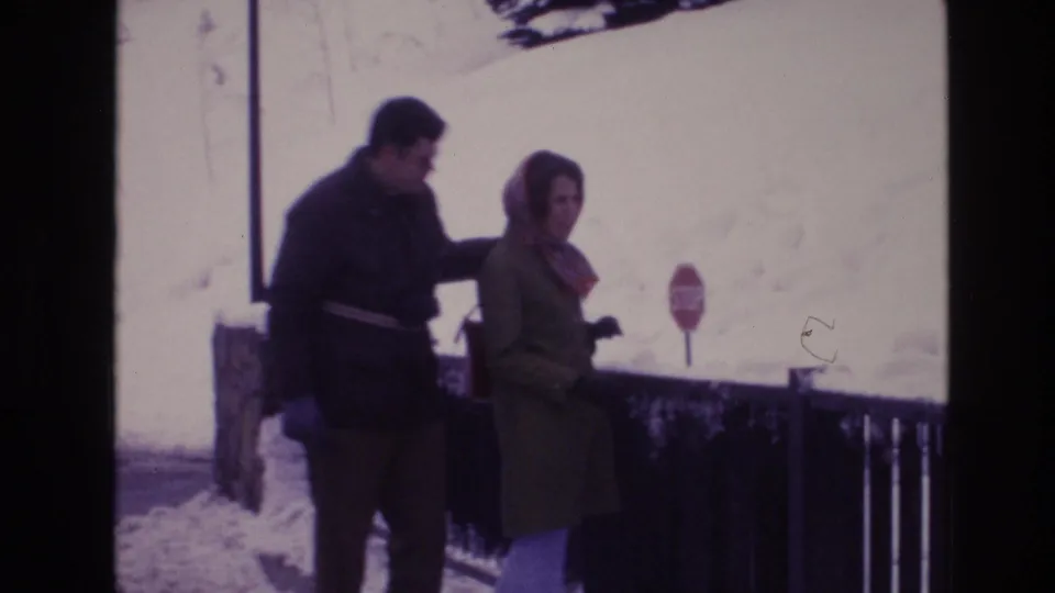 In a 1974 Vail winter resort scene, two adults in coats appear near a railing with snowbanks, a stop sign, and a bright mountain backdrop.