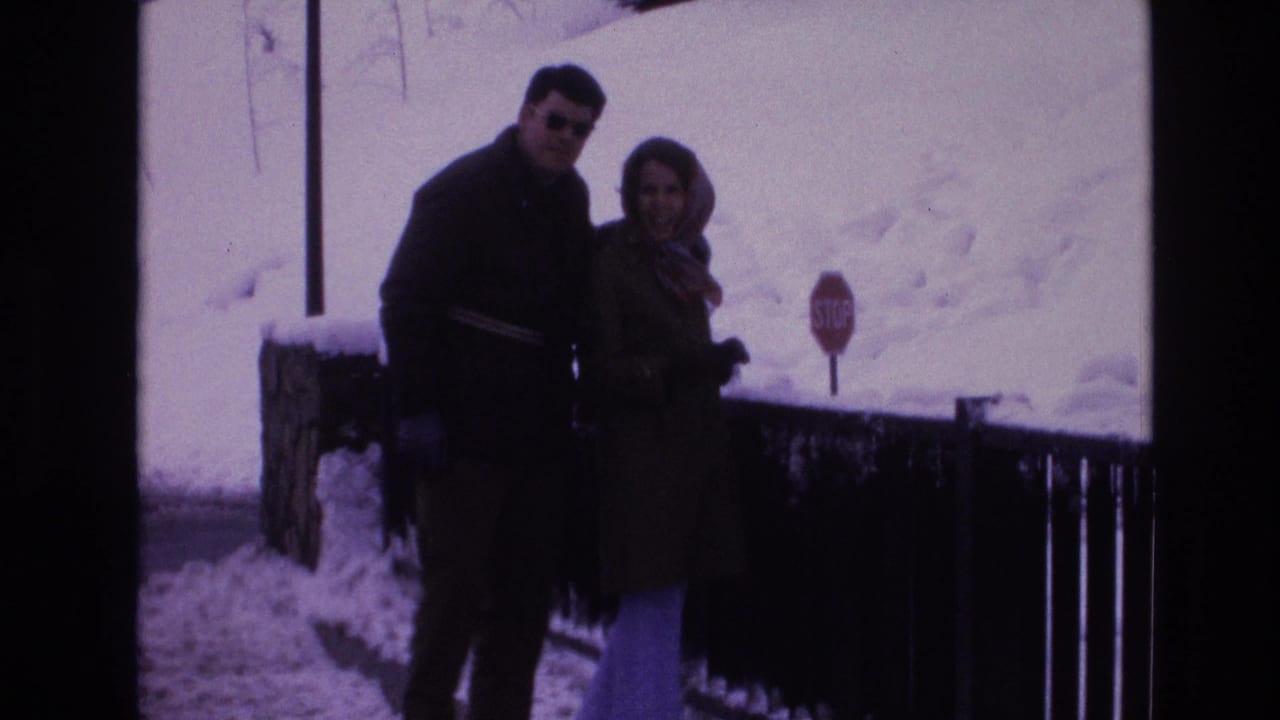 Two adults in winter clothing pose outdoors in snowy Vail, Colorado, with a railing and red stop sign behind them.