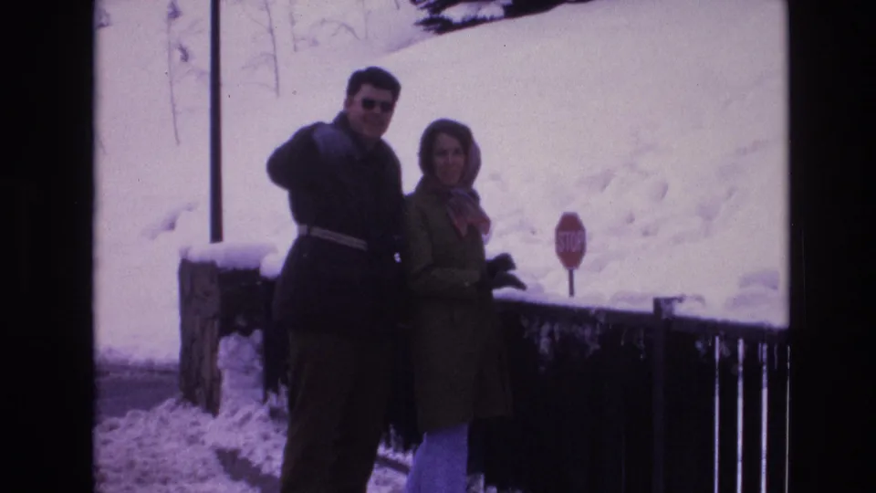 A man in sunglasses and a woman in a headscarf stand close together in a snowy outdoor setting in Vail, Colorado, with a stop sign in the background.
