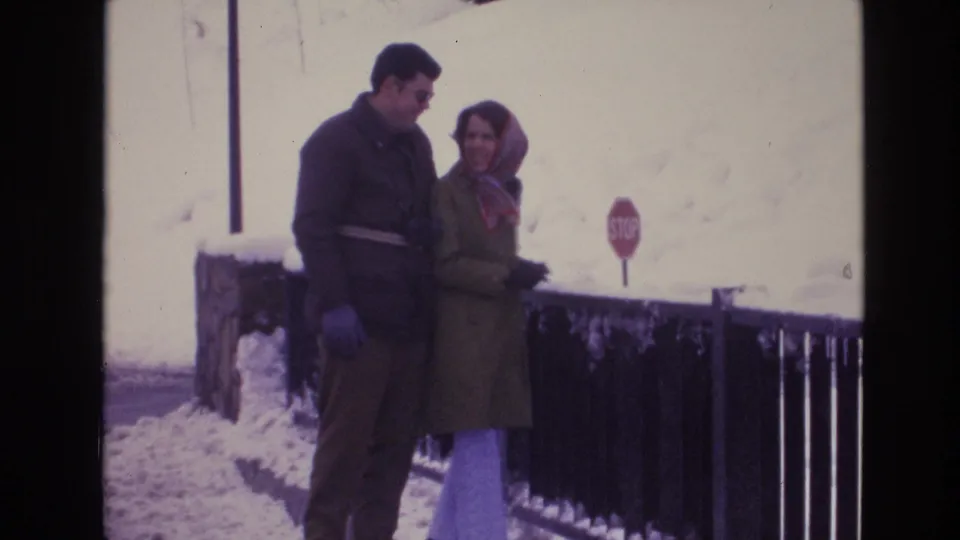 The couple shifts stance and looks toward one another while standing near a snow-covered roadside railing in 1974 Vail, Colorado footage.