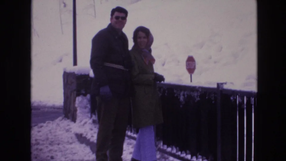 Two bundled adults stand close together in a snowy Vail, Colorado scene, framed by a dark railing and a red stop sign in the background.