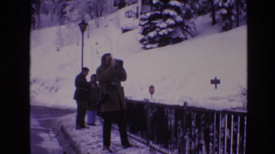 A third adult raises a camera while a couple stands near a snowy railing in Vail, Colorado, with a lamppost, evergreen tree, and stop sign on the hillside behind them.