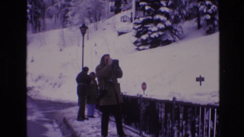 A foreground adult photographs the couple near a snow-covered railing, with a lamppost, red stop sign, evergreen branches, and a white hillside visible in the Vail winter scene.