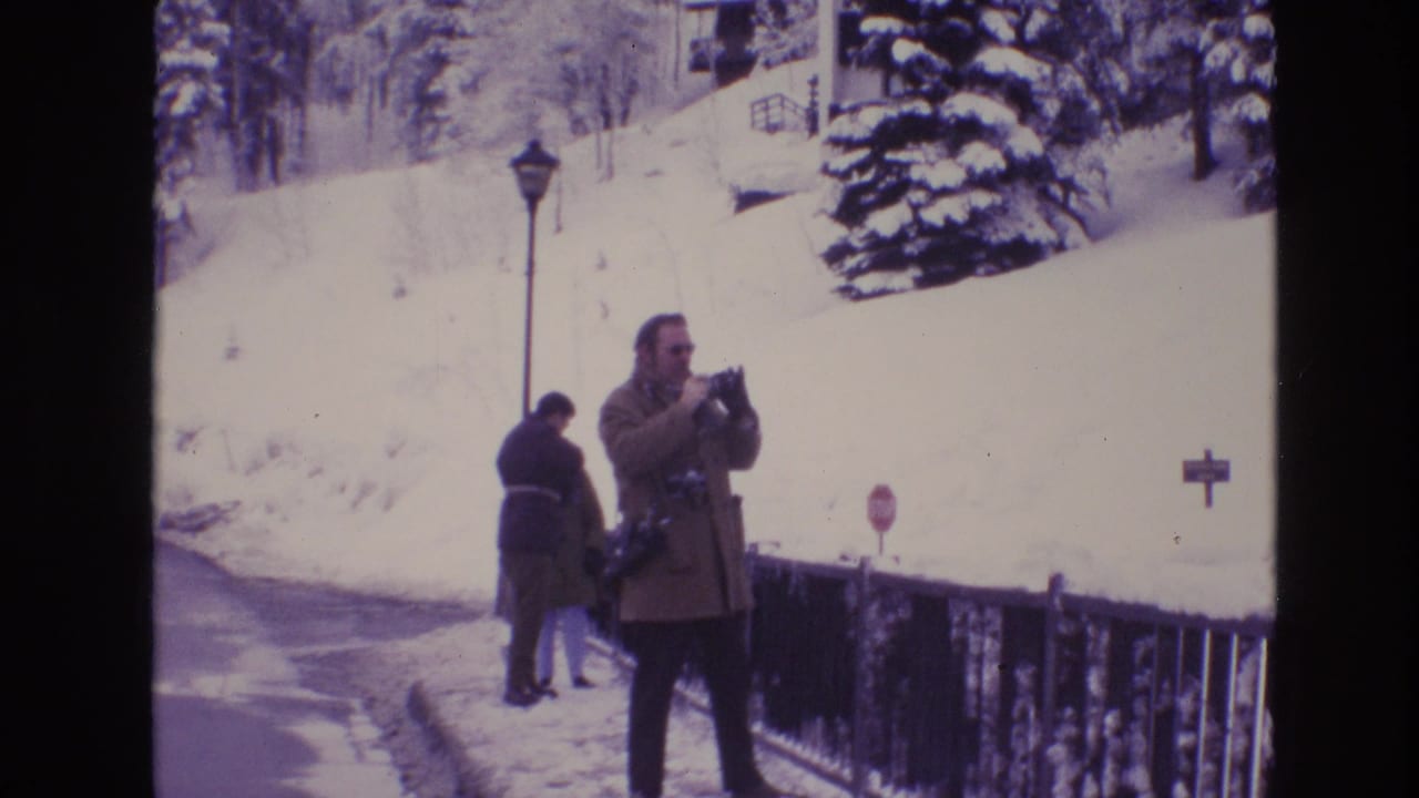 1974 Vail, Colorado home-movie frame showing a man using a still camera on a snow-covered path beside a railing, with two winter-dressed figures near a lamppost in the background.