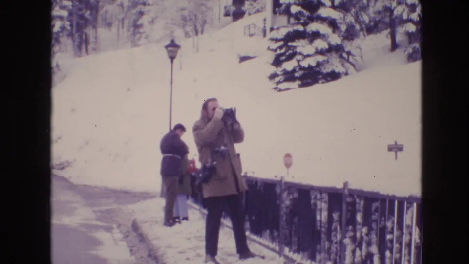 1974 Vail, Colorado frame of a bundled man taking a photograph near a black metal railing, with snowbanks, a lamppost, and two other visitors visible behind him.