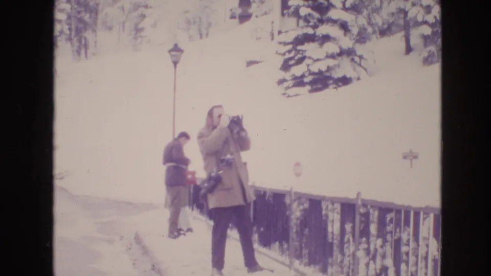 1974 Vail, Colorado home-movie frame showing a man with a camera in the foreground and a second bundled figure bending near the snow along a path bordered by railing and trees.