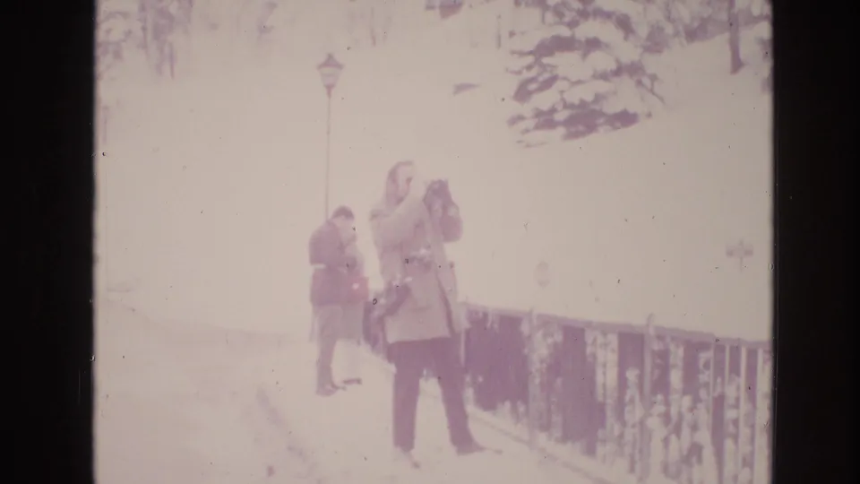 Faded 1974 Vail, Colorado frame with a man holding a camera near a railing, a bundled background figure, and bright snow washing much of the hillside detail.