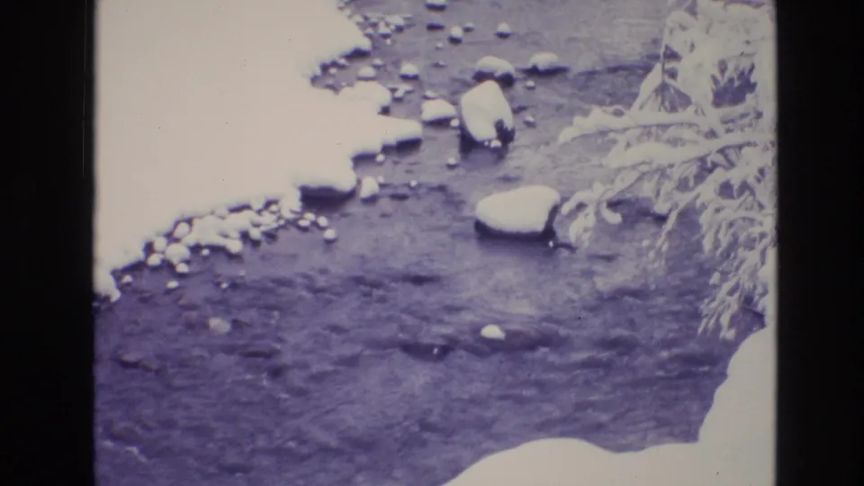 1974 Vail, Colorado 8mm frame looking down at a winter stream with dark moving water, snowbanks, and rounded snow-covered rocks.