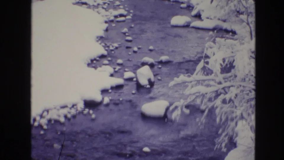 1974 Vail, Colorado 8mm frame showing a partially snow-lined stream with visible stones, dark water, and frosted branches at the bank.
