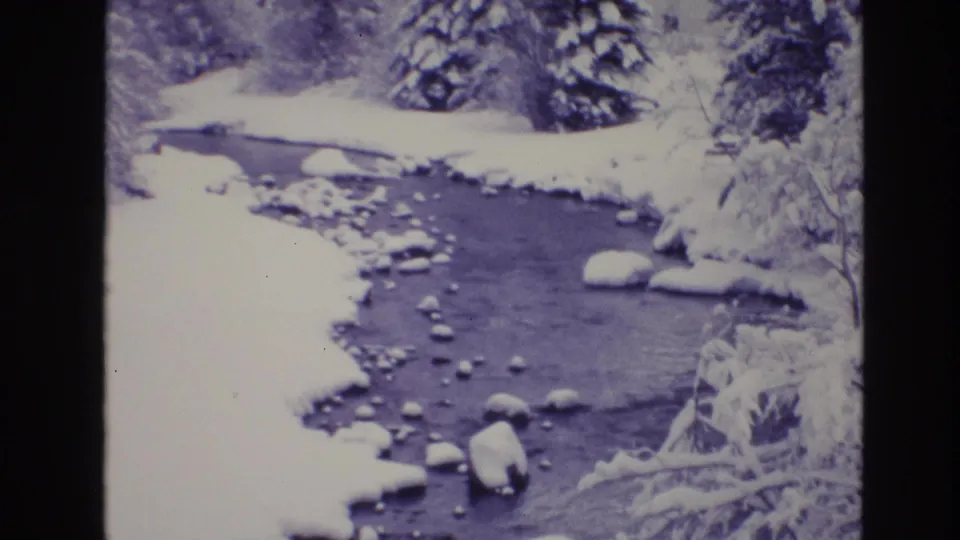 1974 Vail, Colorado 8mm frame of a quiet creek bend bordered by snow, evergreen growth, and several rounded rocks in the water.