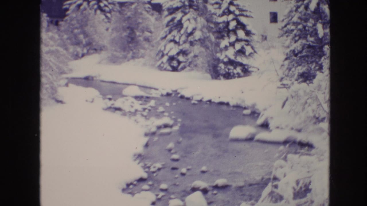 1974 Vail, Colorado 8mm frame showing a snow-covered stream bordered by evergreens, snowbanks, and partially visible alpine buildings.