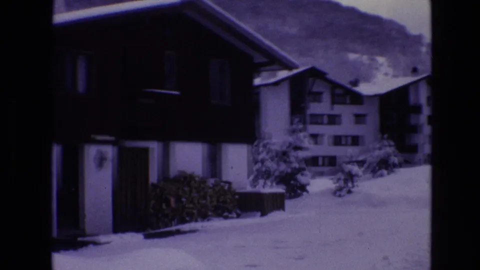 1974 Vail, Colorado 8mm frame showing a dark chalet-like building, snow-covered ground, nearby resort buildings, and a mountain slope in the background.