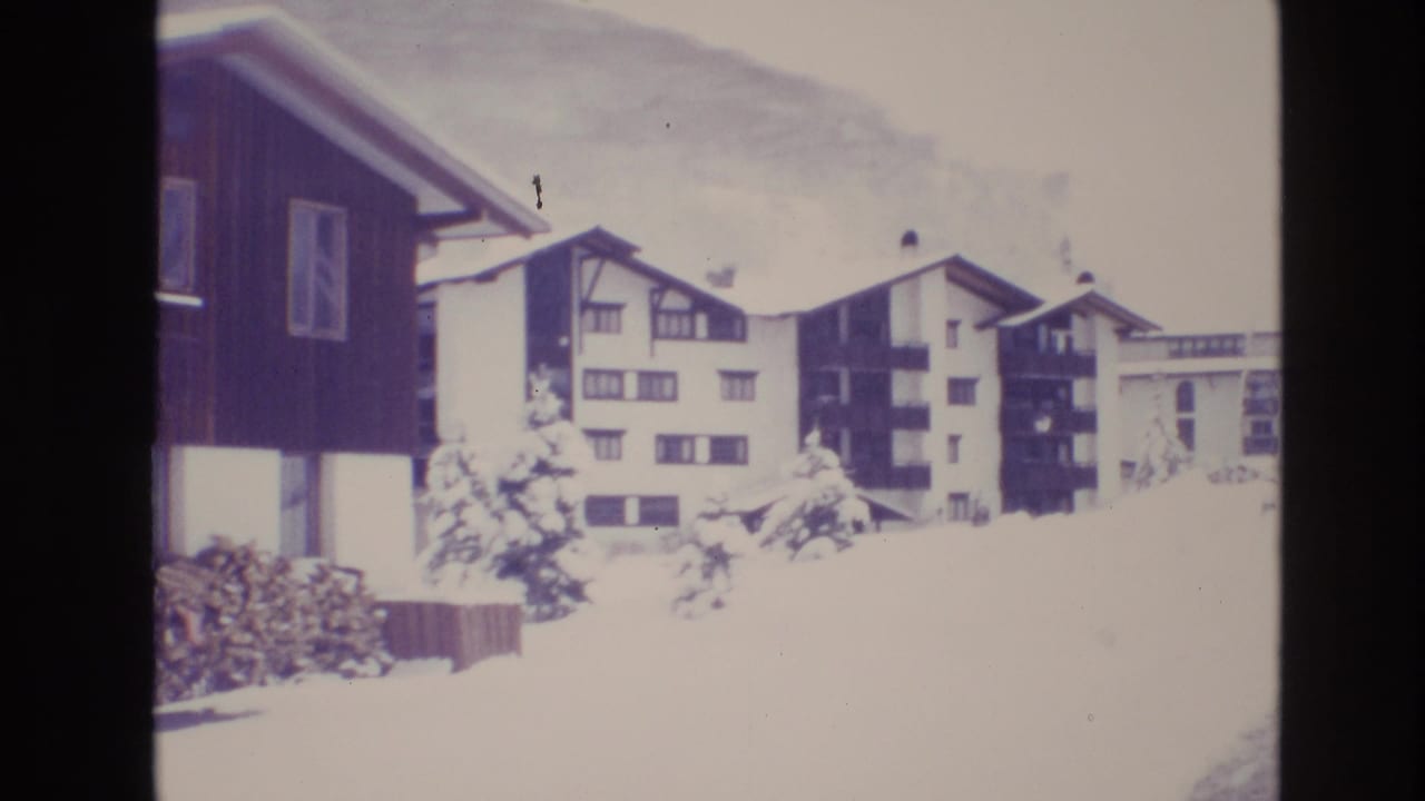 Snowy resort apartment buildings with balconies, evergreens, and mountain background.