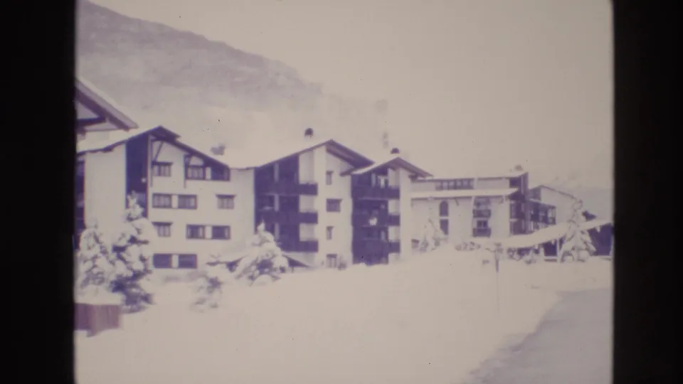 1974 Vail, Colorado home-movie frame showing multiple resort apartment buildings, snow-covered evergreens, a broad snowfield, and a cleared edge at lower right.