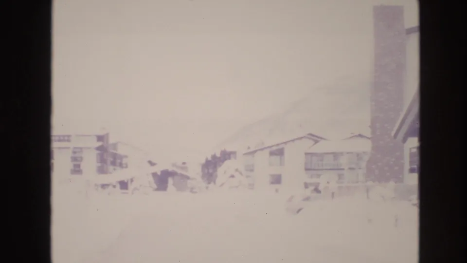 1974 Vail home-movie frame showing snow-covered resort lodging, balconies, pitched roofs, and a tall chimney-like vertical structure.