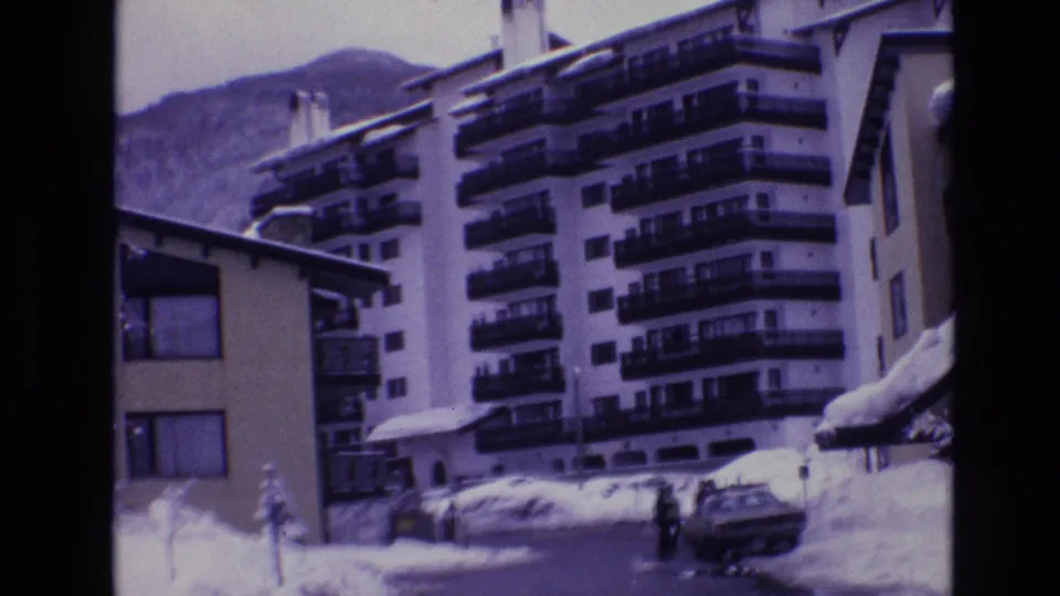 1974 Vail 8mm frame of a multi-story resort building with balconies, snowbanks, parked cars, and distant people near the entrance or street.