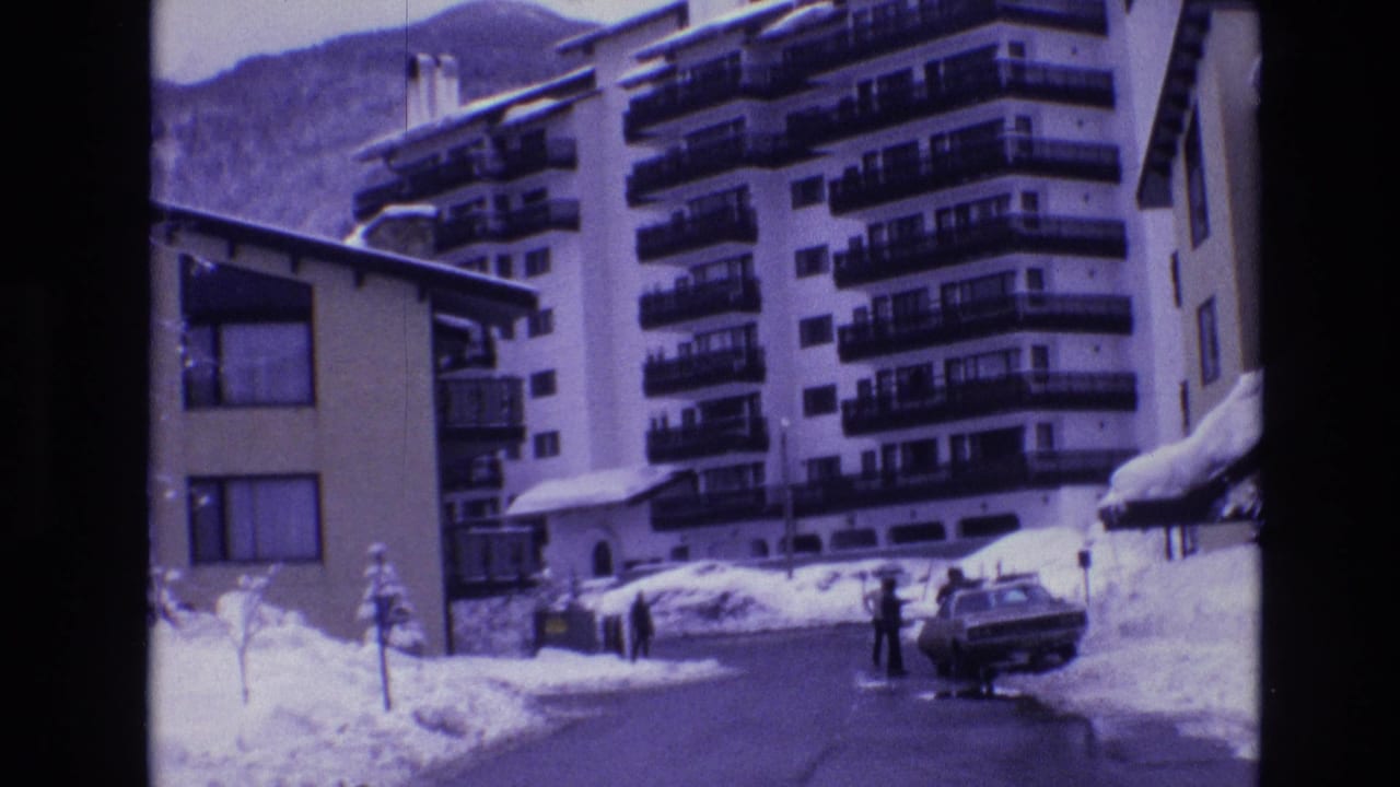 1974 Vail, Colorado home-movie frame showing a snow-lined resort street, a parked sedan, distant pedestrians, multi-story hotel balconies, and a mountain backdrop.