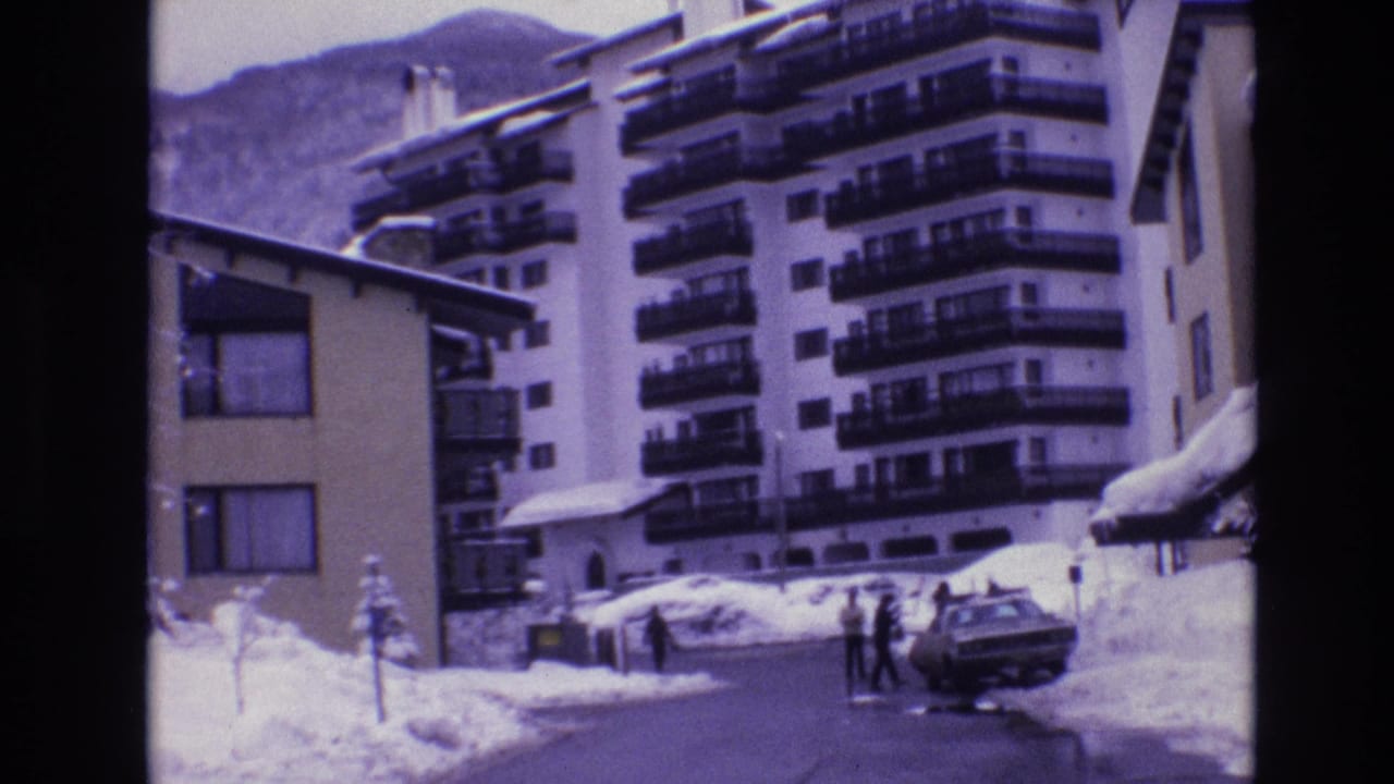People walk near a parked car on a snowy street below resort hotel balconies.