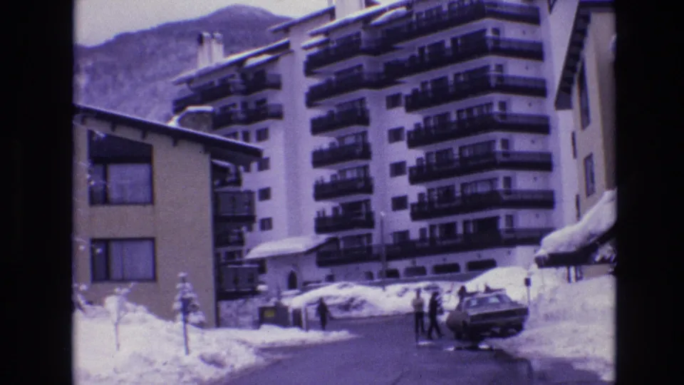 1974 Vail, Colorado frame with pedestrians crossing or standing near a parked sedan on a snowy street in front of resort lodging and mountain scenery.