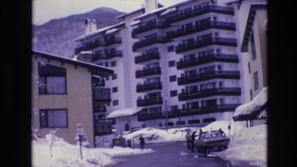1974 Vail, Colorado home-movie frame showing several distant people by a parked sedan on a snow-covered resort street with balcony-fronted lodging behind them.