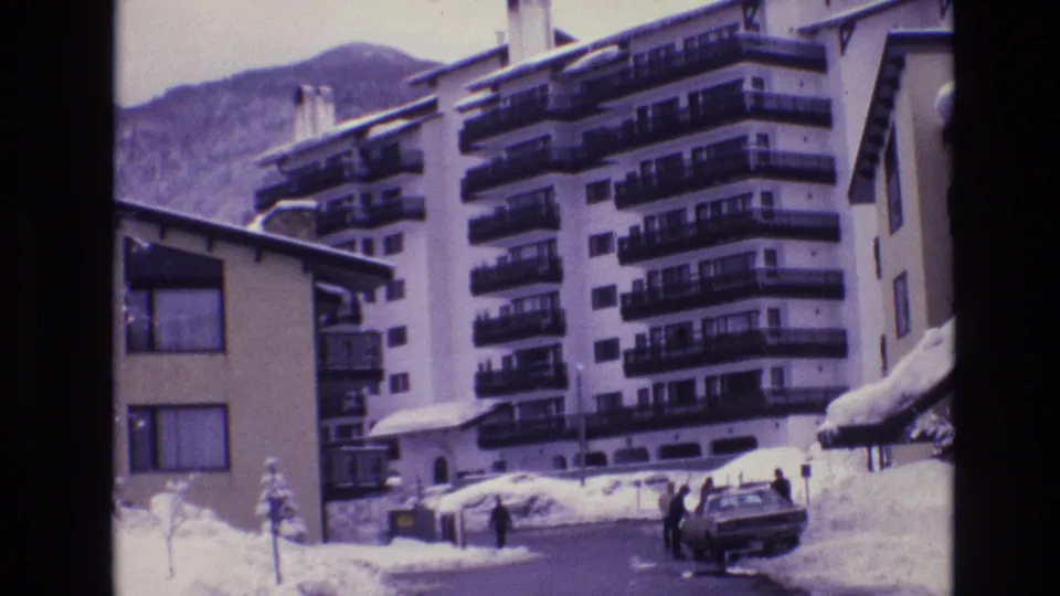 1974 Vail, Colorado frame showing people gathered near a parked sedan on a snow-lined road, with multi-story resort buildings, balconies, and a mountain behind them.