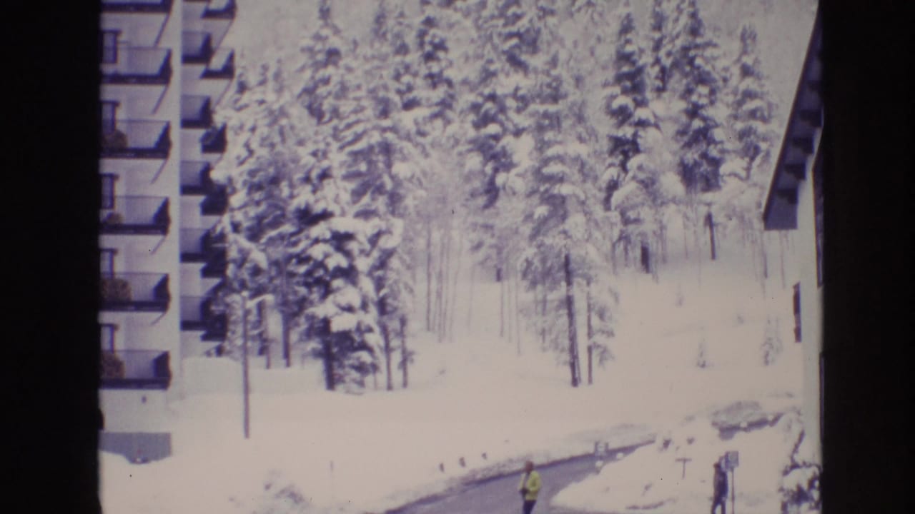 1974 Vail, Colorado 8mm frame showing a snow-covered apartment area, tall pine trees, balconies at left, and distant pedestrians near a plowed winter road.