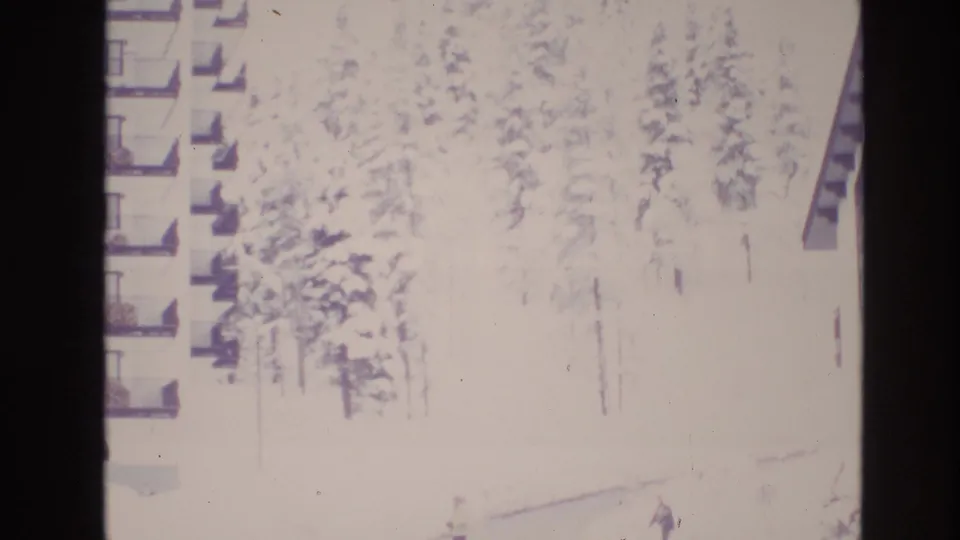 Overexposed 8mm winter frame from Vail, Colorado showing apartment balconies, dense snow-laden pines, and a small distant pedestrian in the snow.