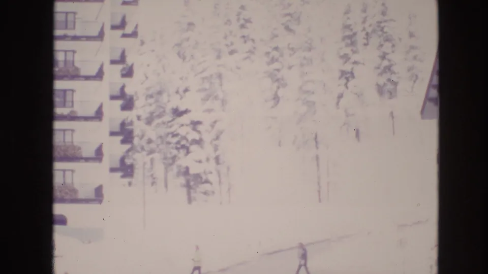 1974 Vail, Colorado home-movie frame with apartment balconies at left, snowy pines behind, and a bundled pedestrian crossing the overexposed winter foreground.