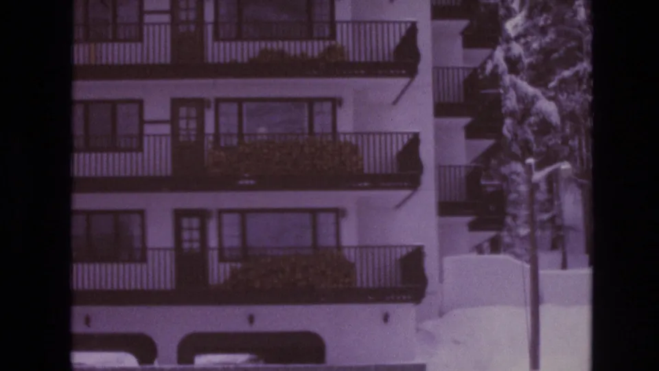 Closer 8mm view of a Vail apartment exterior in winter, with stacked balconies, dark railings, shrubs, snowbanks, and pine trees along the building.