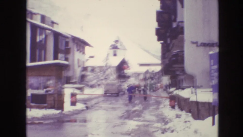 1974 Vail, Colorado home-movie view down a snowy village street with chalet-style buildings, parked cars, and small distant pedestrians.