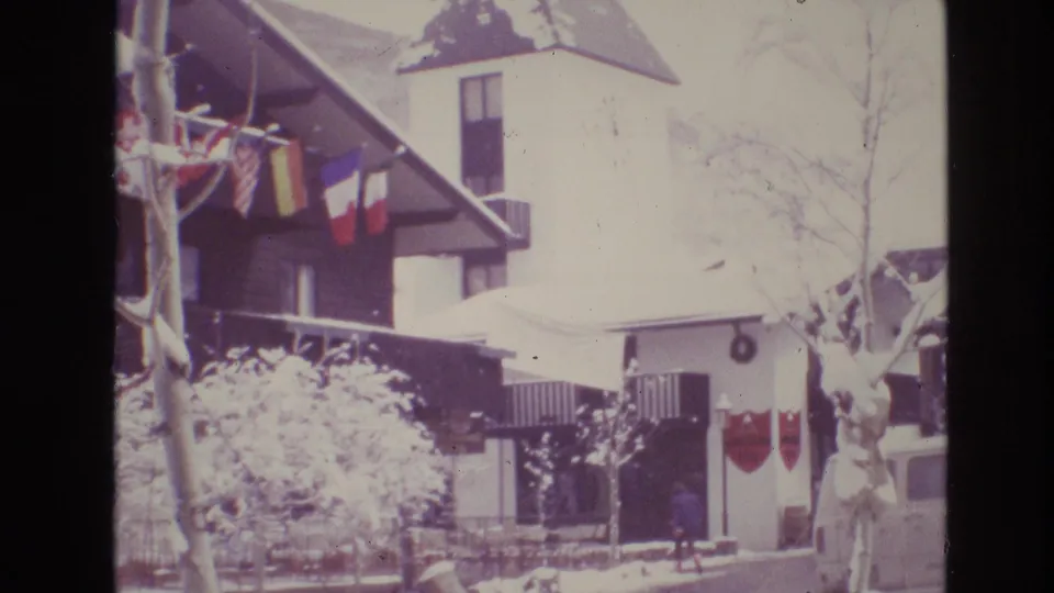1974 Vail, Colorado frame showing snow-covered chalet-style buildings, hanging flags, a tower structure, shrubs, and small distant people near the walkway.