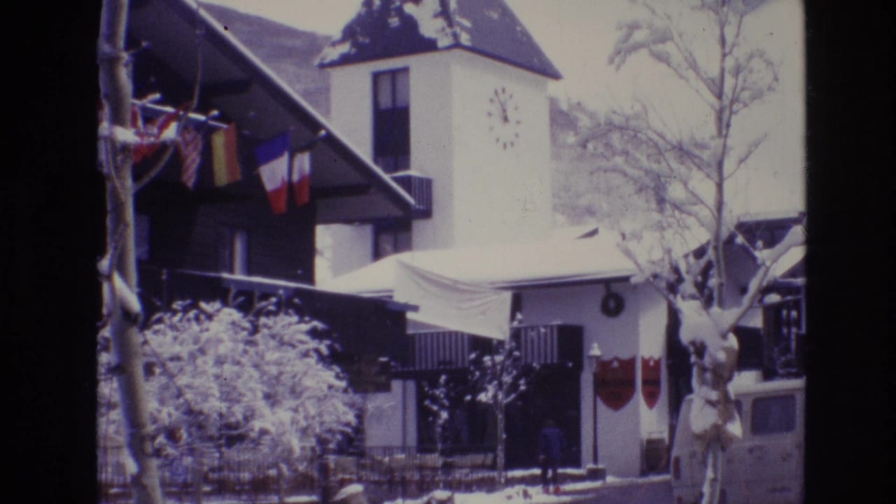 1974 Vail, Colorado 8mm frame showing a snow-covered resort hotel exterior with international flags, a clock tower, bare trees, and a white van near the entrance.