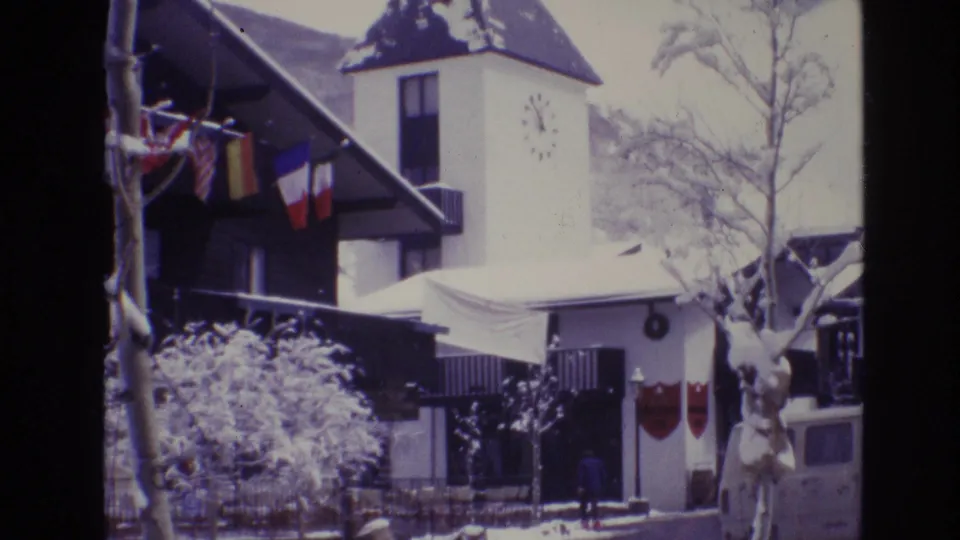 1974 Vail, Colorado 8mm frame showing a snow-covered resort hotel exterior with international flags, a clock tower, bare trees, and a white van near the entrance.