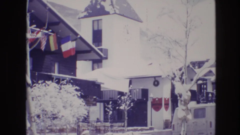 1974 Vail, Colorado frame of a snow-covered resort building with international flags, a clock tower, bare trees, a visible entrance, and a white van parked nearby.