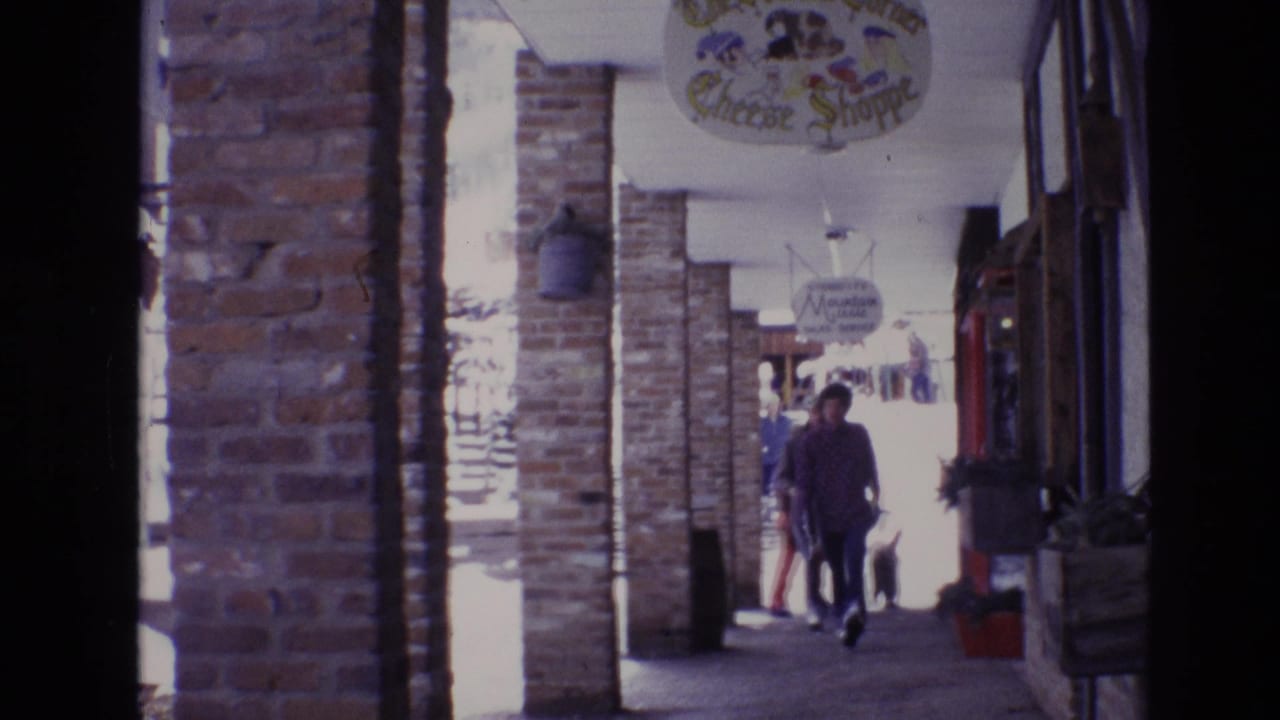 1974 Vail, Colorado 8mm frame showing a covered brick arcade with pedestrians moving past storefronts and hanging shop signs in a snowy ski-resort plaza.