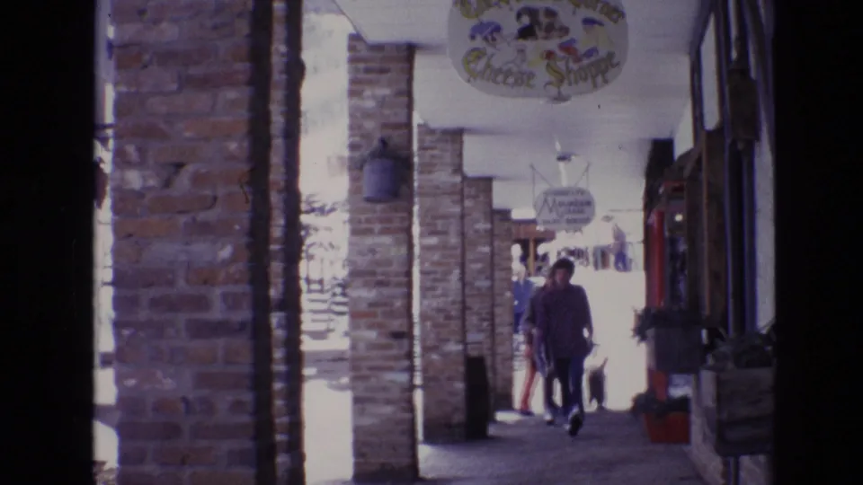 1974 Vail, Colorado 8mm frame showing a covered brick arcade with pedestrians moving past storefronts and hanging shop signs in a snowy ski-resort plaza.