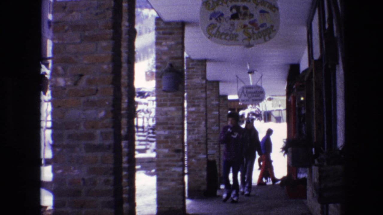 1974 Vail, Colorado frame of bundled pedestrians walking through a shaded covered shopping arcade, with brick columns and shop signage visible overhead.