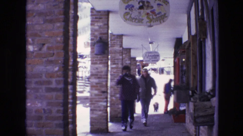 1974 Vail, Colorado 8mm frame showing visitors and a dog in a covered brick shopping arcade, with the Cheese Shoppe sign and another hanging sign visible above.