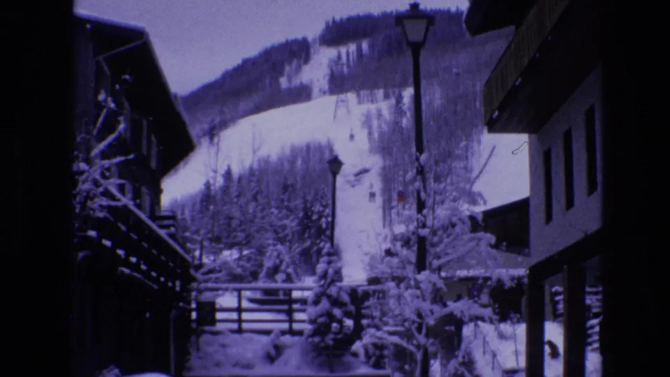 1974 Vail, Colorado frame looking from the resort village toward snow-covered buildings, lamp posts, trees, and a visible ski trail on the mountain slope.