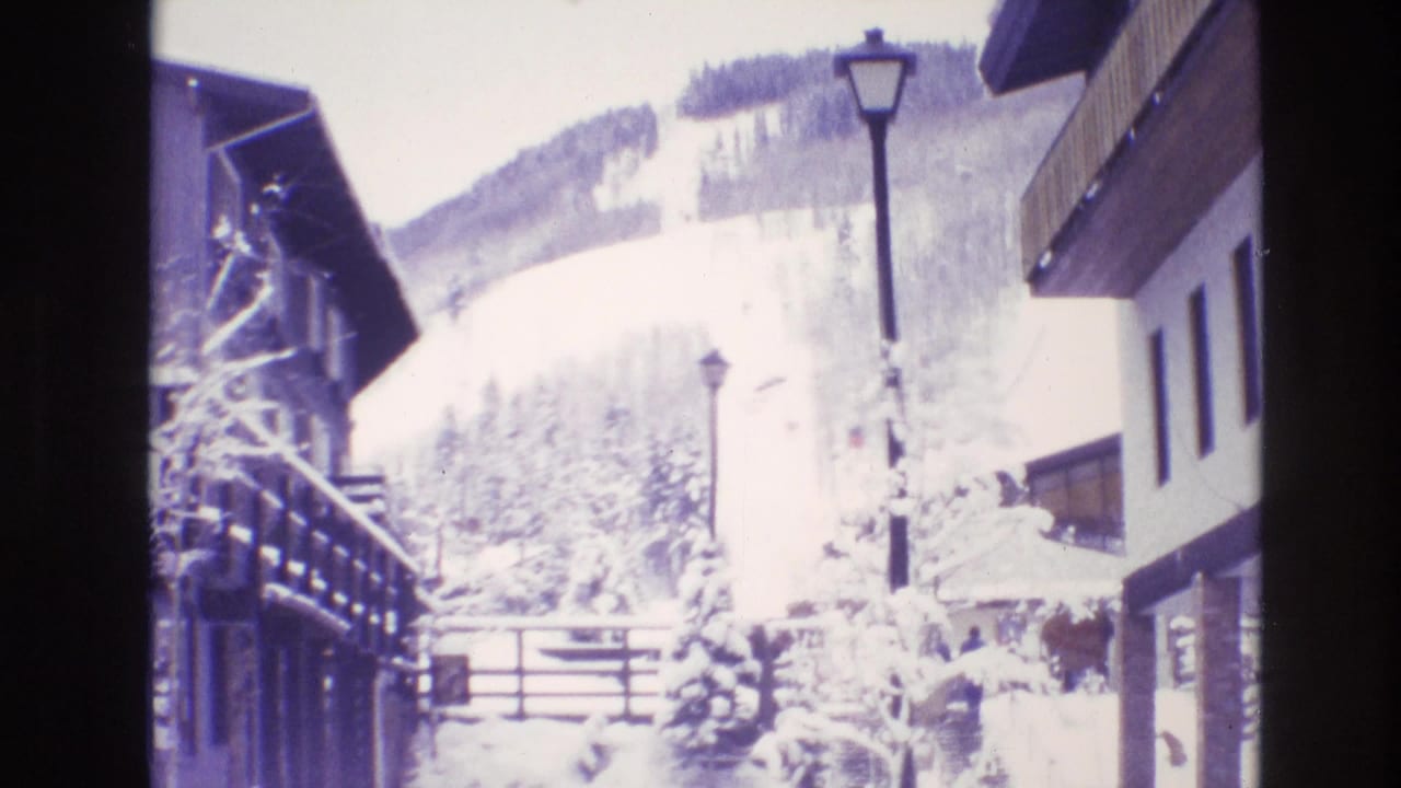 1974 Vail, Colorado 8mm frame showing a snow-covered resort lane between lodge buildings, with a dark light pole, fence, trees, and a ski slope beyond.