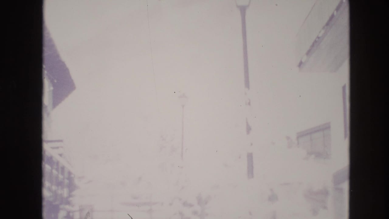 Overexposed snowy street with faint lodge walls, lamp posts, and building outlines.