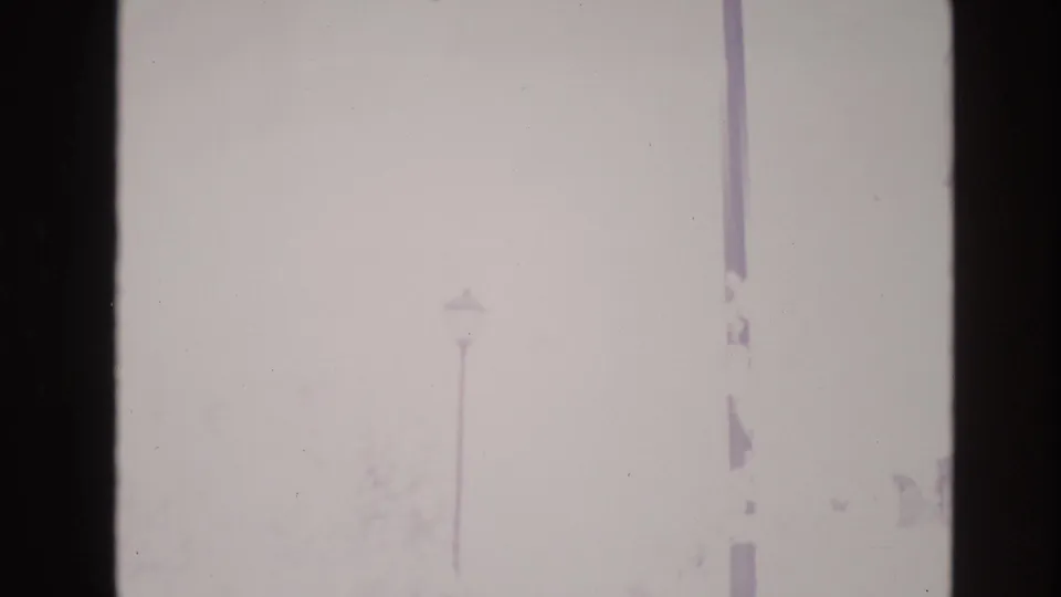 1974 Vail 8mm frame reduced to a pale whiteout view, with only faint pole shapes and a shadowed building edge remaining visible.