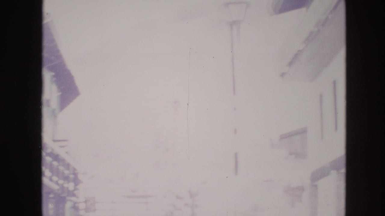 Hazy overexposed 8mm frame of a snowy Vail, Colorado street in 1974, with building edges and a tall lamp post barely visible through bright winter light.