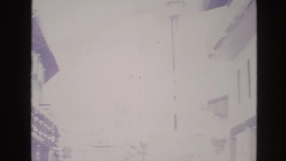 Hazy overexposed 8mm frame of a snowy Vail, Colorado street in 1974, with building edges and a tall lamp post barely visible through bright winter light.