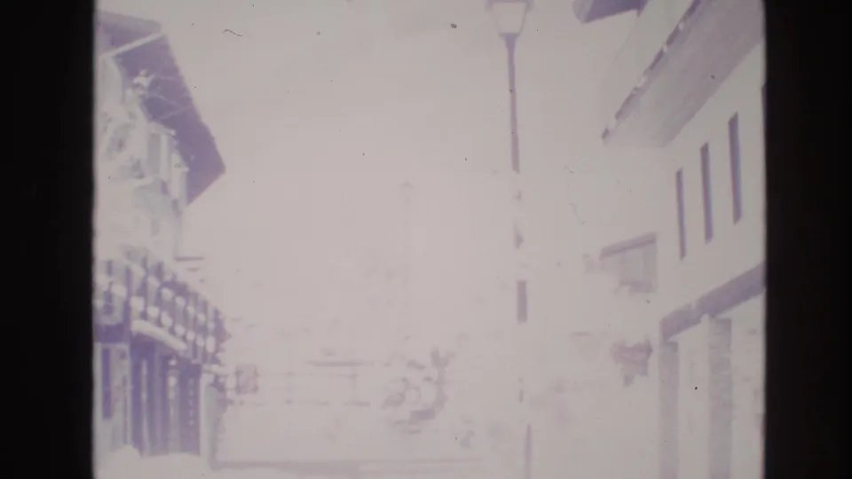 Overexposed 1974 home-movie view of a snowy Vail street, showing wooden building fronts, balconies, and a lamp post in washed winter daylight.