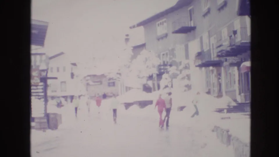 1974 8mm frame of pedestrians in winter clothing walking through a snowy Vail street lined with wooden storefronts, balconies, awnings, and a signboard.